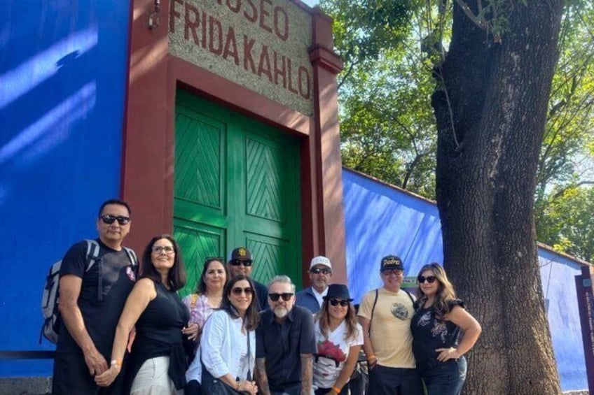 A group of people posing in front of a building with a green door and a sign that reads 'FRIDAKAHLO.' 1