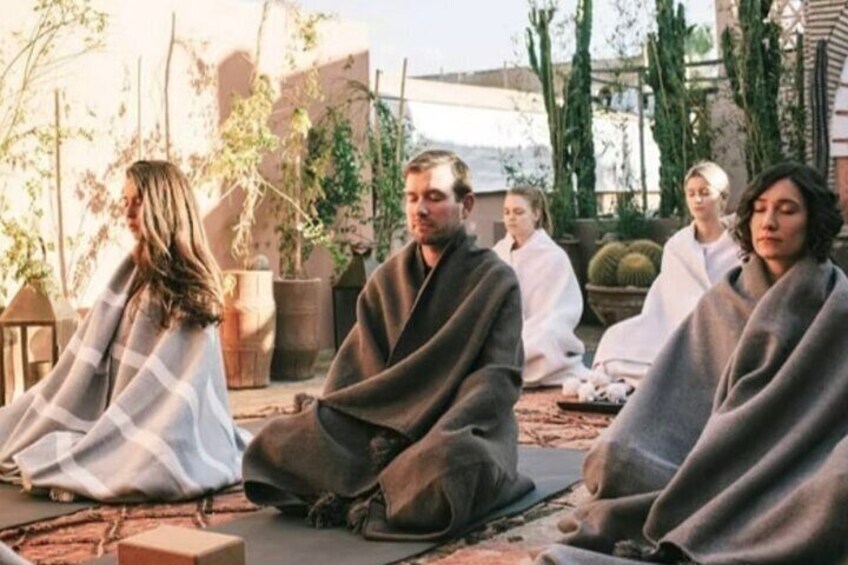 Eine Gruppe von Menschen sitzt draußen im Schneidersitz, eingehüllt in Decken, in entspannter Atmosphäre. Im Hintergrund sind Kakteen und Topfpflanzen zu sehen. 1