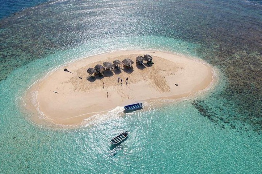 An aerial view of a small island with clear turquoise waters, a sandy beach, and a few thatched-roof structures. 1