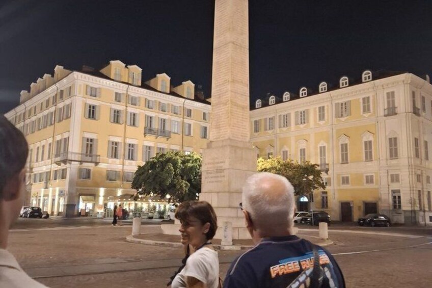A nighttime city square with illuminated buildings, a central monument, and a few people. 1