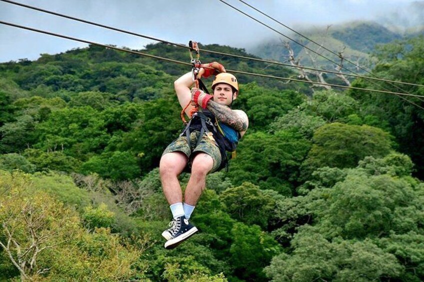 A person zip-lining through a forested area with dense green foliage and hills in the background. 1
