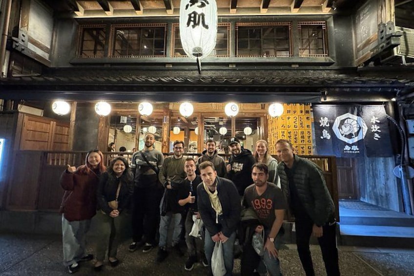 Un grupo de personas posando frente a un edificio tradicional con arquitectura de madera y letreros iluminados. 1