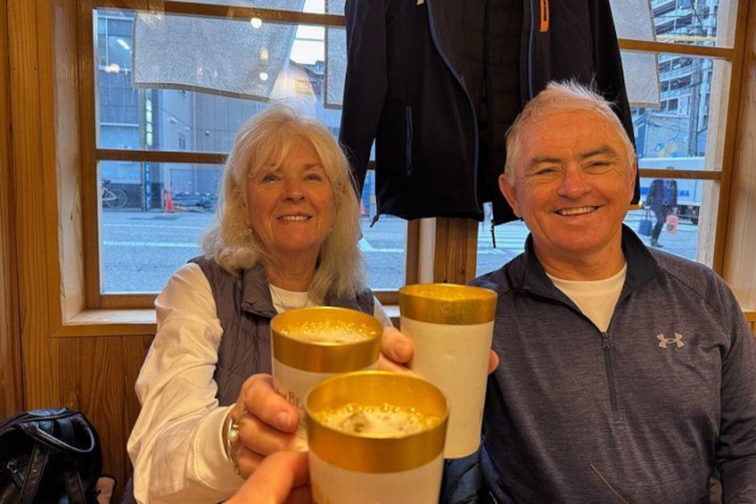 Two people holding mugs with a frothy beverage, a window with a view of a street, and a black jacket hanging on the wall. 3