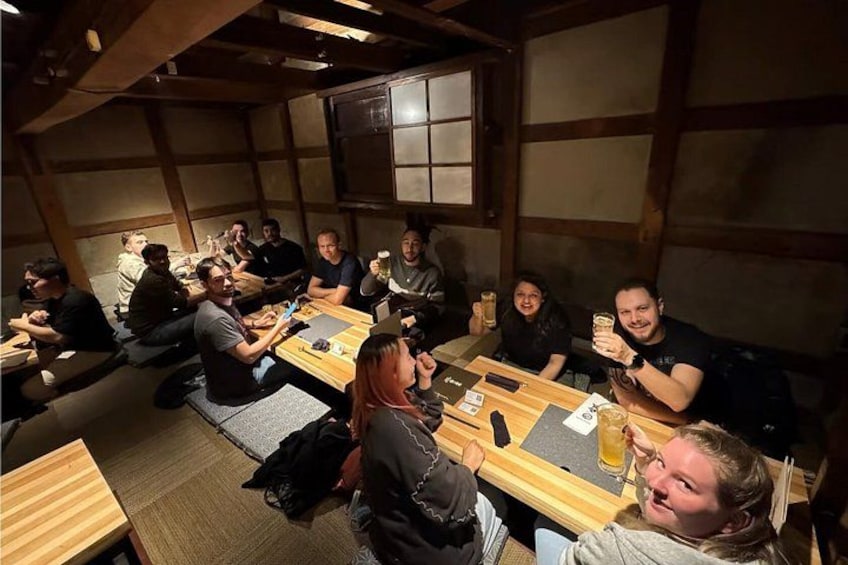 Un grupo de personas sentadas alrededor de una mesa de madera en una habitación con esteras de tatami, conversando y sosteniendo bebidas. 3