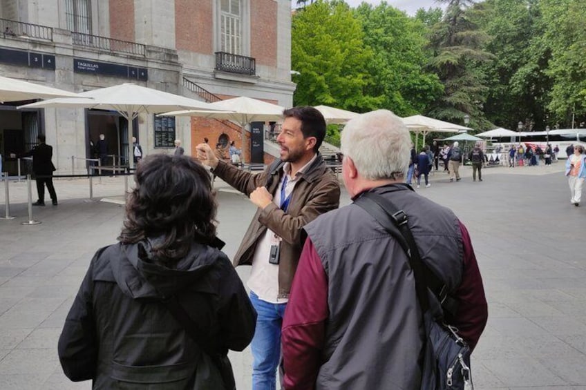 Un groupe de personnes se tient à l’extérieur, peut-être sur une place publique ou dans un parvis, avec des bâtiments et des arbres en arrière-plan. 2
