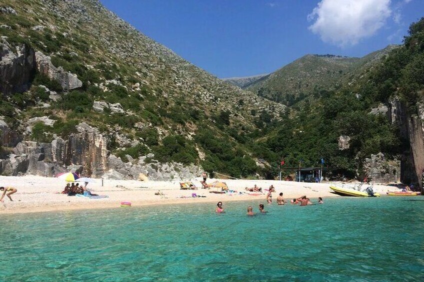 Ein Strand mit klarem türkisblauem Wasser, Menschen beim Schwimmen und Sonnenbaden, und felsige Hügel im Hintergrund. 5