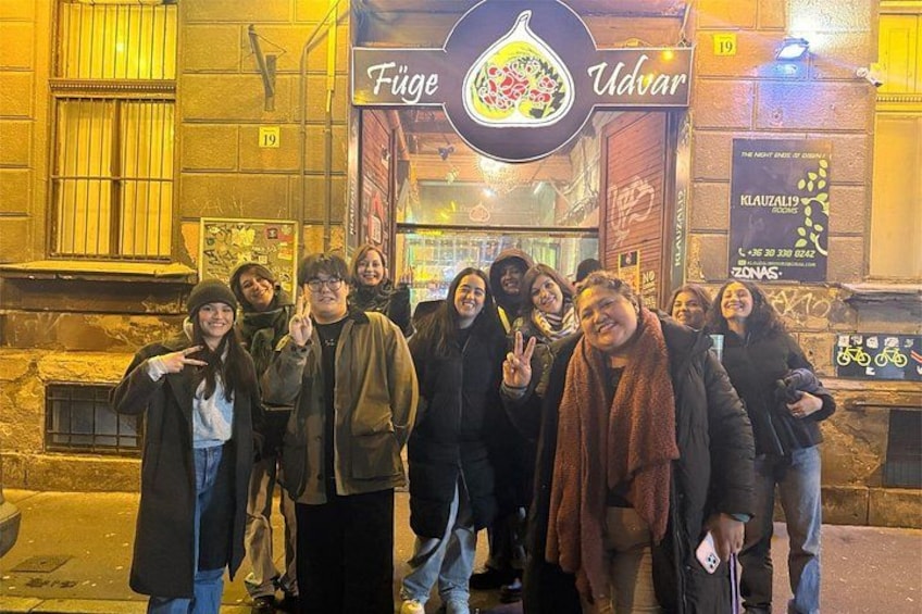 Un grupo de personas posando frente a un restaurante llamado 'Füge Udvar', con un letrero y una señal de estacionamiento para bicicletas al fondo. 2
