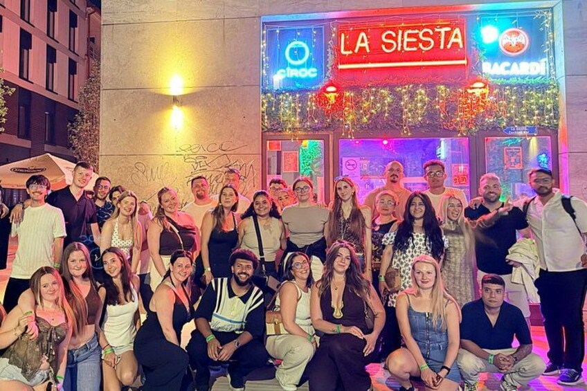 A group of people posing in front of a brightly lit bar named 'LA SIERRA' with neon signs and outdoor seating. 1