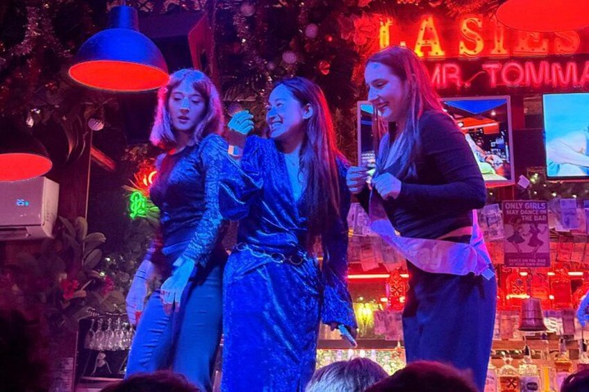 A stage with three people, a neon sign, and a bar in the background. 3