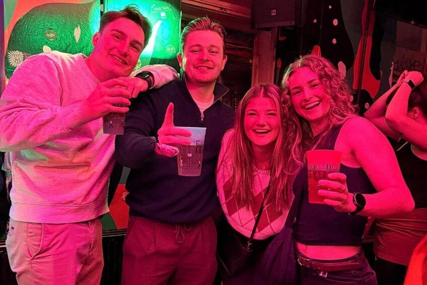 A group of people in a dimly lit indoor setting, possibly a bar or club, with a green neon sign in the background. 2