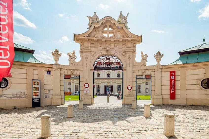 An ornate entrance with a sign for tickets, a cobblestone courtyard, and a building in the background. 2