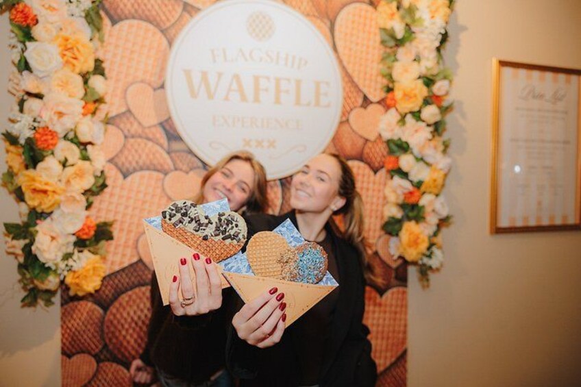 Two people holding waffle boxes in front of a decorated backdrop with a waffle theme. 2