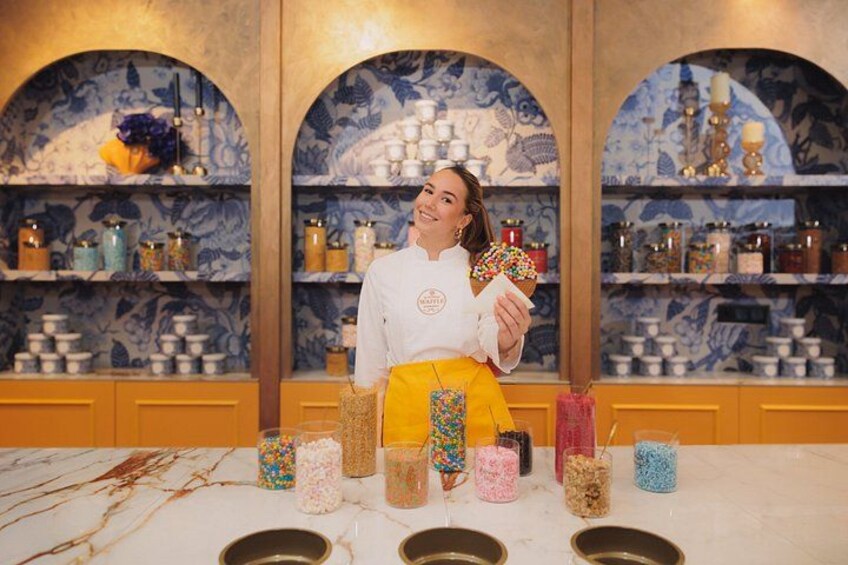 A person in a white chef's coat holding a colorful object, with shelves of jars and containers in the background. 1