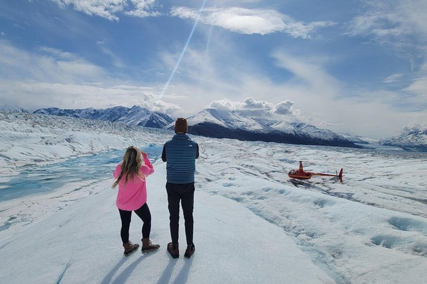 A snowy landscape with a person and a child standing, a helicopter in the background, and mountains in the distance. 3