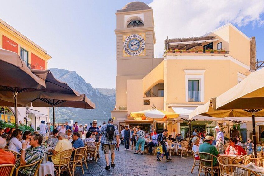 A bustling outdoor café with wicker chairs and umbrellas, a clock tower, and a mountain backdrop. 2
