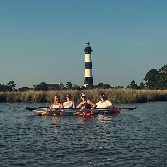 Four people rowing a boat on a calm body of water with a lighthouse in the background. 3