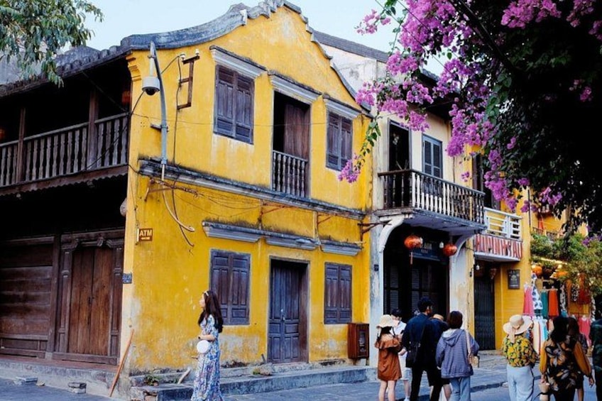 A yellow building with a balcony, a sign that reads 'ATM,' and people walking by. 1