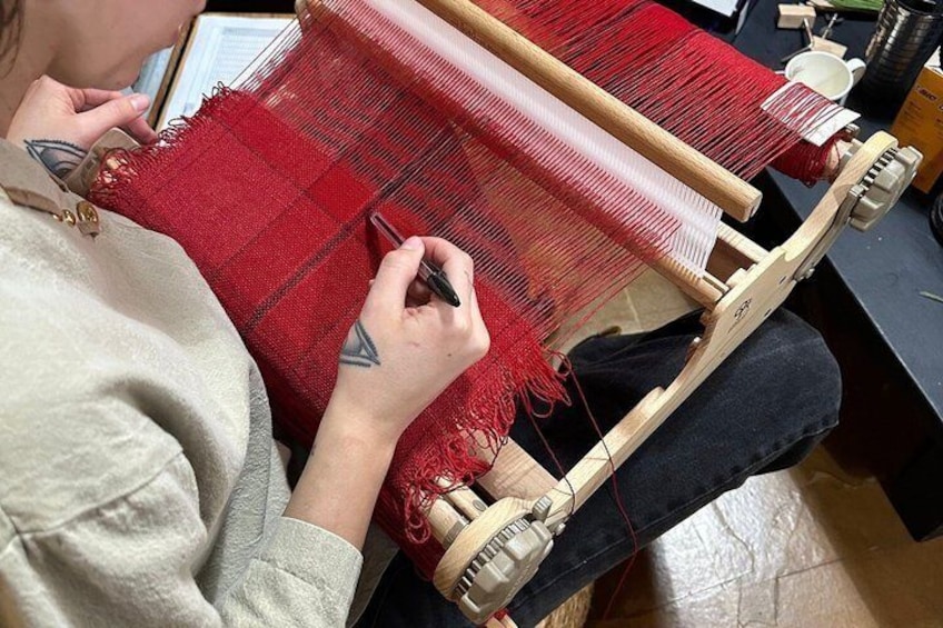 Una persona tejiendo en un telar de madera con hilos rojos y blancos. 3