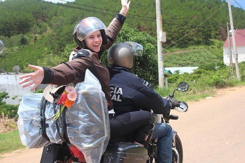 Two people on a motorcycle, one wearing a helmet and the other with arms outstretched, on a rural road with greenery in the background. 1