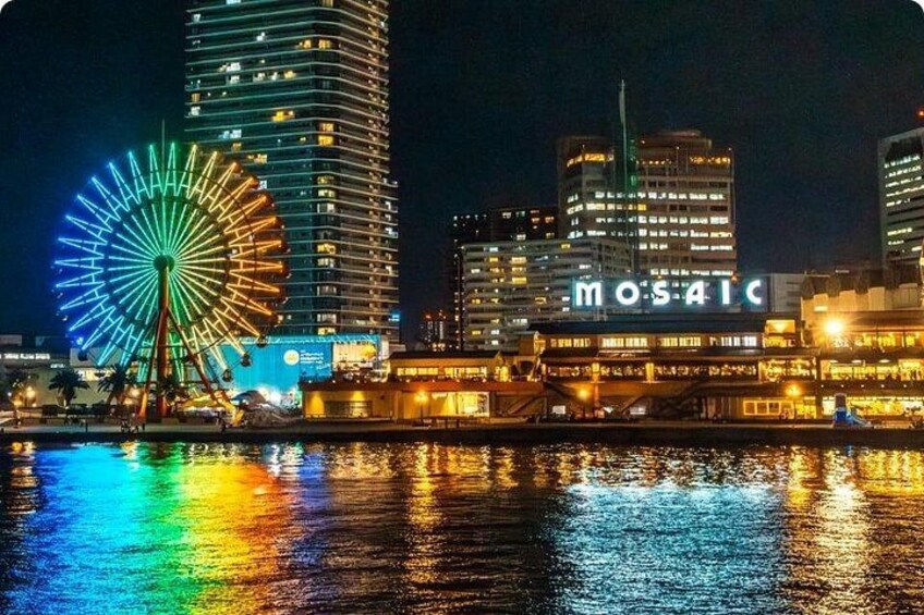 Ein belebtes Stadtbild bei Nacht mit einem hell erleuchteten Riesenrad und einem Gebäude, das 'MOSAIC' anzeigt. 2