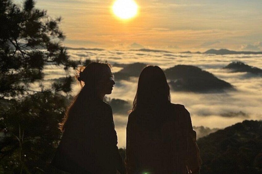 Deux personnes sont assises sur une colline, d’où elles dominent un paysage montagneux, avec un coucher de soleil en arrière-plan. 2