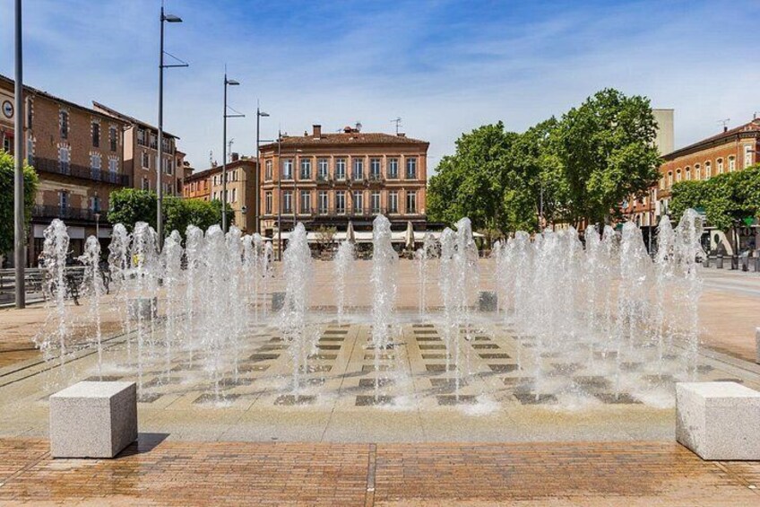 Ein Brunnen mit Wasserstrahlen auf einem Platz, im Hintergrund Gebäude. 3