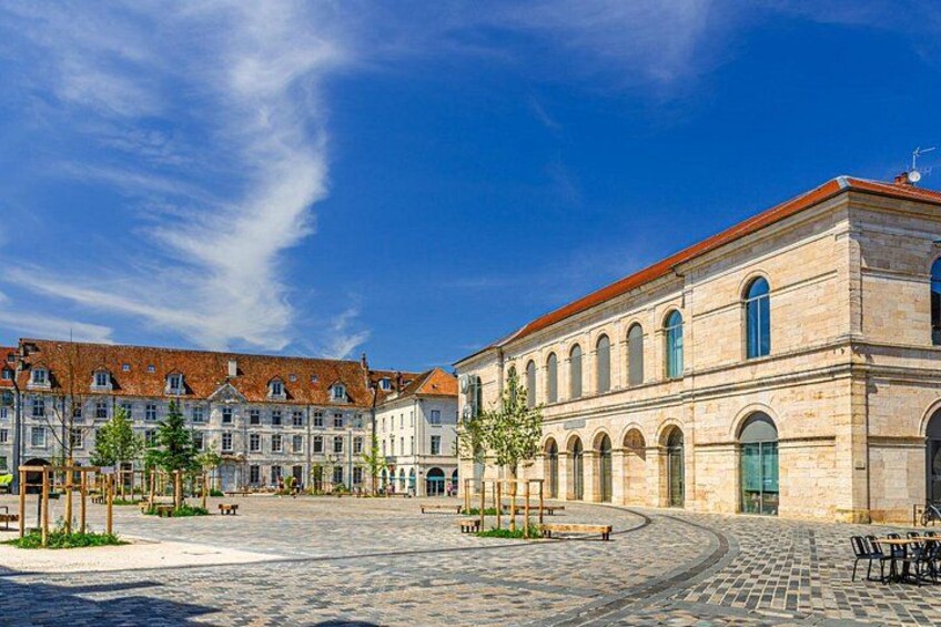 Ett torg med kullerstenar, omgivet av historiska byggnader, bänkar och träd. 2