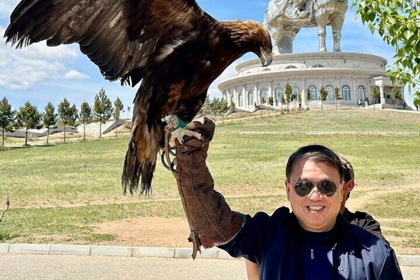 Une personne tenant un grand oiseau de proie, peut-être un aigle, avec une statue et un bâtiment en arrière-plan. 2