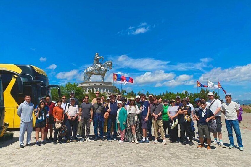 A group of people standing in front of a monument with a statue, a bus, and flags. 2
