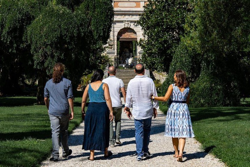 En grupp människor promenerar på en grusgång i en park med träd och en byggnad i bakgrunden. 5