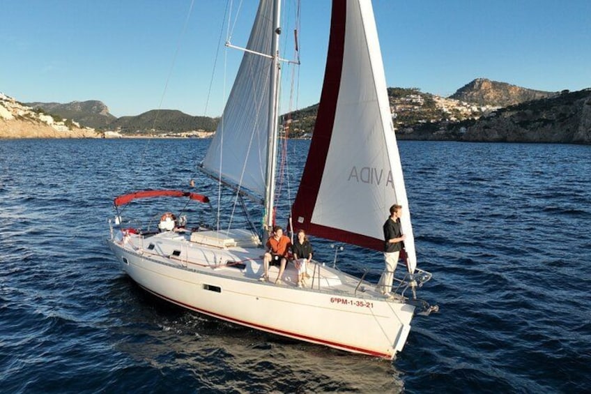A sailboat with a white hull and red accents, sailing on calm waters with a coastal landscape in the background. 1