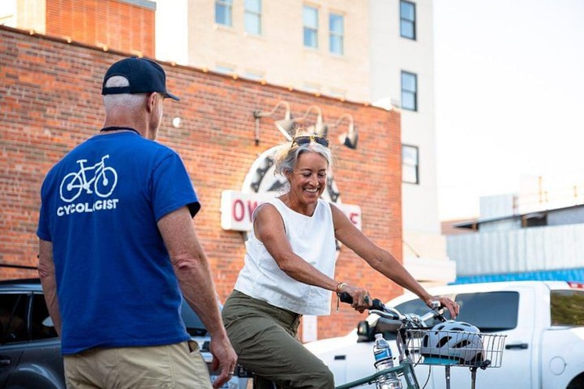 A person riding a bicycle with a basket, a man in a blue shirt, and a brick building in the background. 2