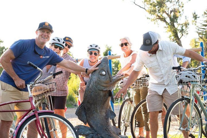 A group of people on bicycles, a fish sculpture, and trees in the background. 4