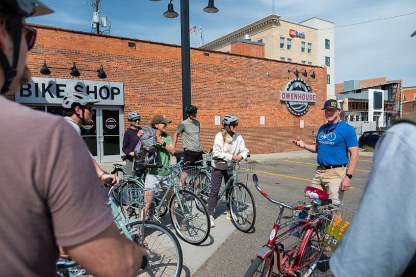 A group of people with bicycles in front of a brick building with a bike shop sign. 2