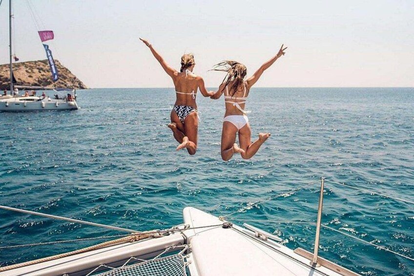 Two people jumping off a boat into the sea, with a sailboat and rocky island in the background. 1