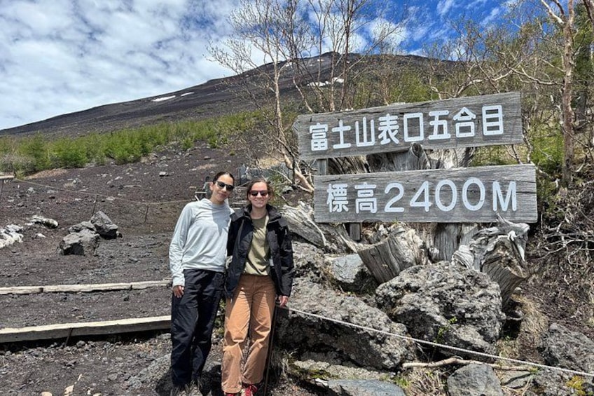 Dos personas de pie frente a un letrero con texto japonés y un paisaje montañoso al fondo. 5