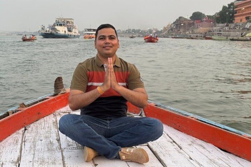 A person sitting cross-legged on a boat with a river and boats in the background. 1