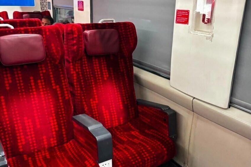 A red patterned seat with a grey armrest and a window with a red emergency exit sign. 2
