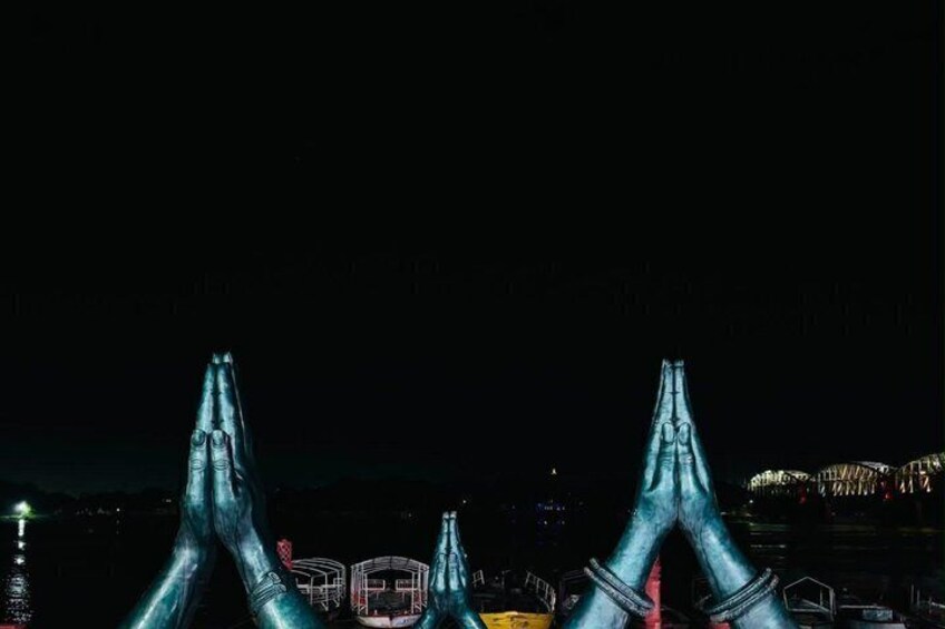 A night scene with illuminated structures resembling praying hands, a cityscape in the background, and a faintly visible bridge. 3