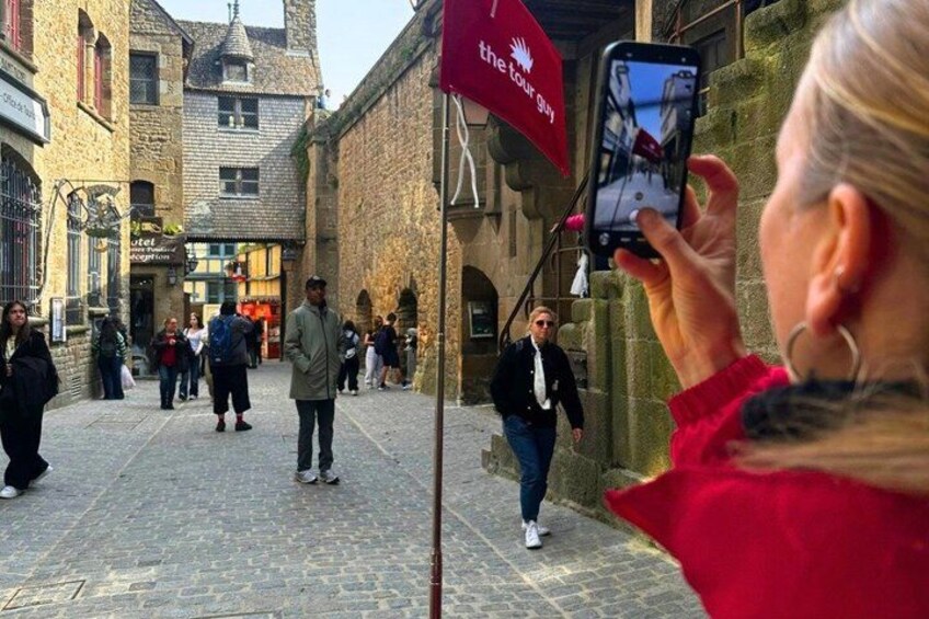 A cobblestone street with people walking, a red flag with white text, and a person taking a photo. 3
