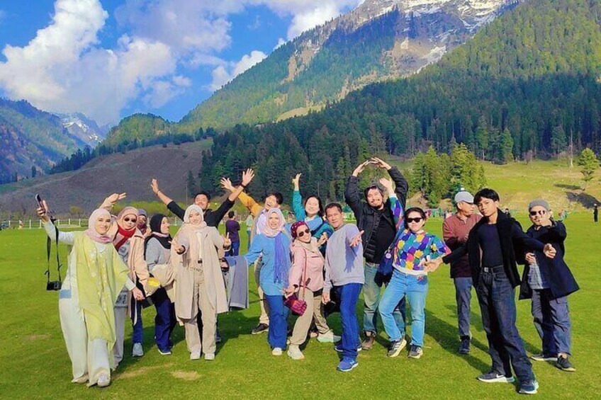A group of people posing in a grassy field with mountains in the background. 1
