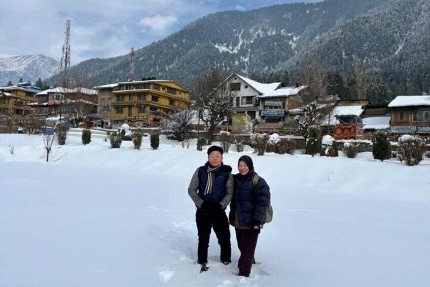 A couple standing in a snowy village with buildings and mountains in the background. 3