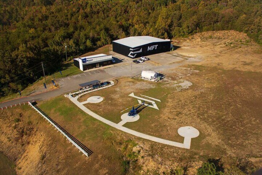 An aerial view of a facility with a large building labeled 'MFT', a helipad, and a parking area surrounded by a forested landscape. 4