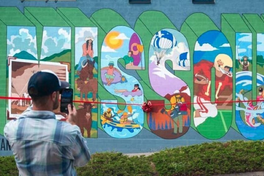 A mural with various colorful scenes and a person taking a photo. 1