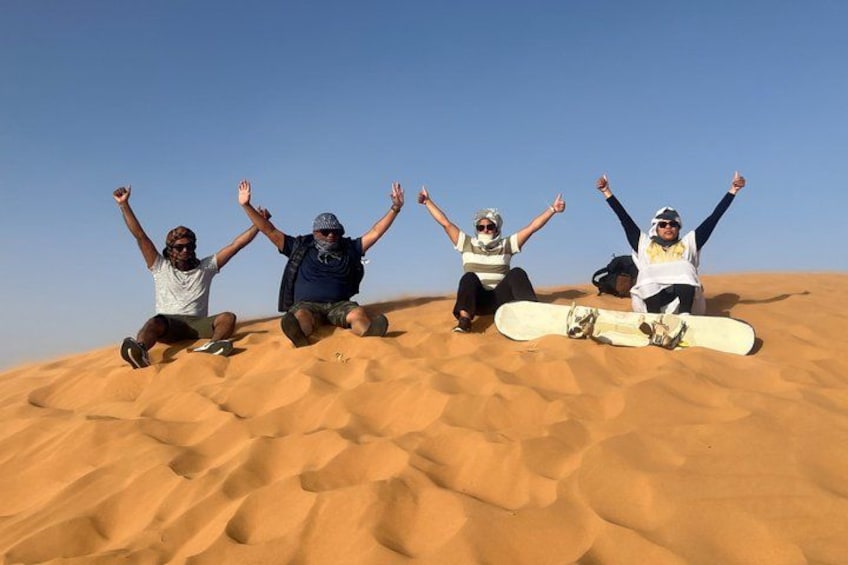 Four people sitting on a sand dune with a snowboard in front of them. 5