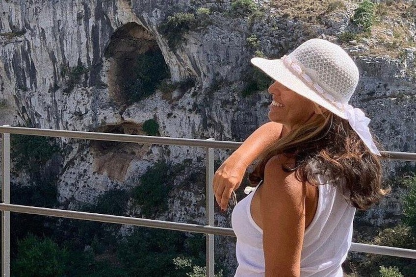 A person wearing a white hat and white top, standing near a railing with a rocky cliff in the background. 4