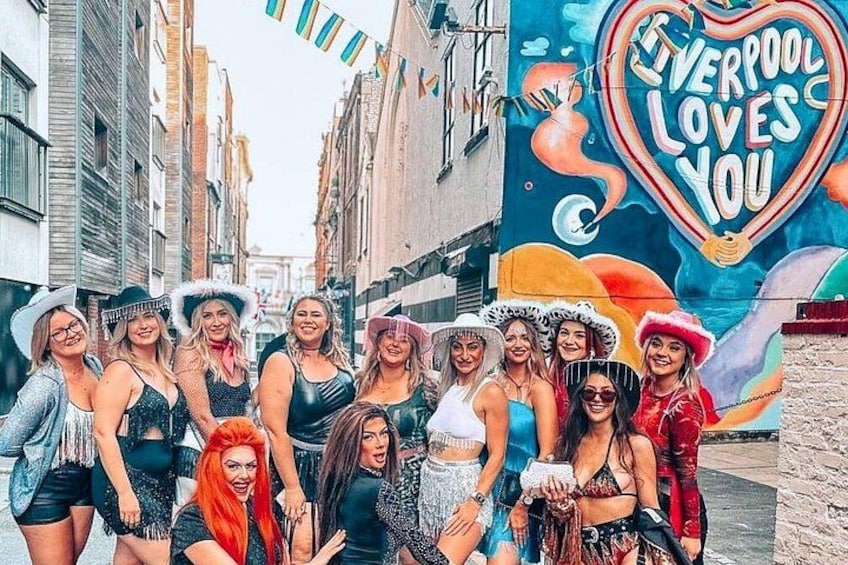 Un grupo de personas posando frente a un mural con el texto 'Liverpool Loves You' y un símbolo de corazón. 2