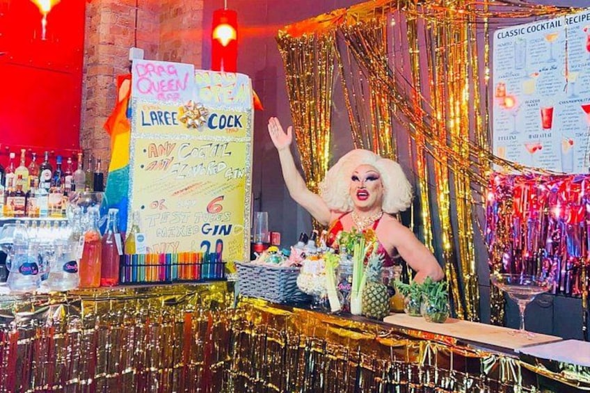 Una persona vestida de reina del drag en un bar, con un cartel que anuncia un cóctel de drag. 3