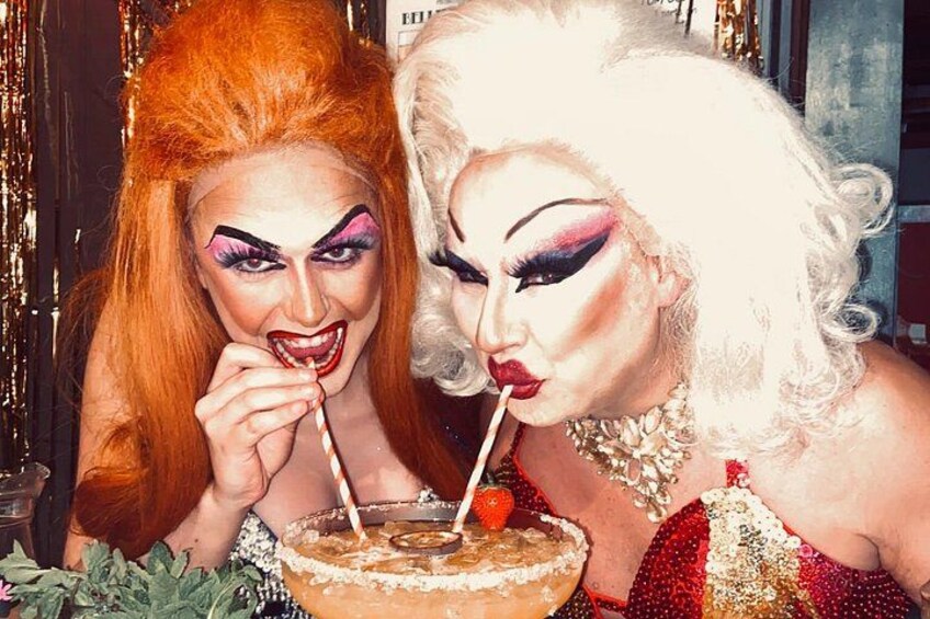 Two individuals with elaborate makeup and costumes, one with red hair and the other with blonde hair, holding a drink with a straw. 2