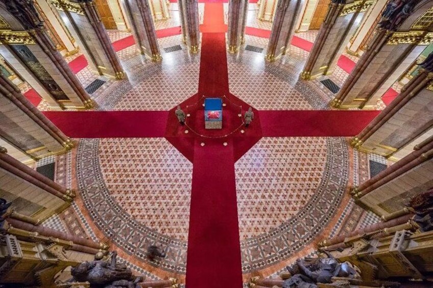 Un salón imponente con una alfombra roja que conduce a una plataforma central, columnas ornamentadas y baldosas de suelo con diseños. 4